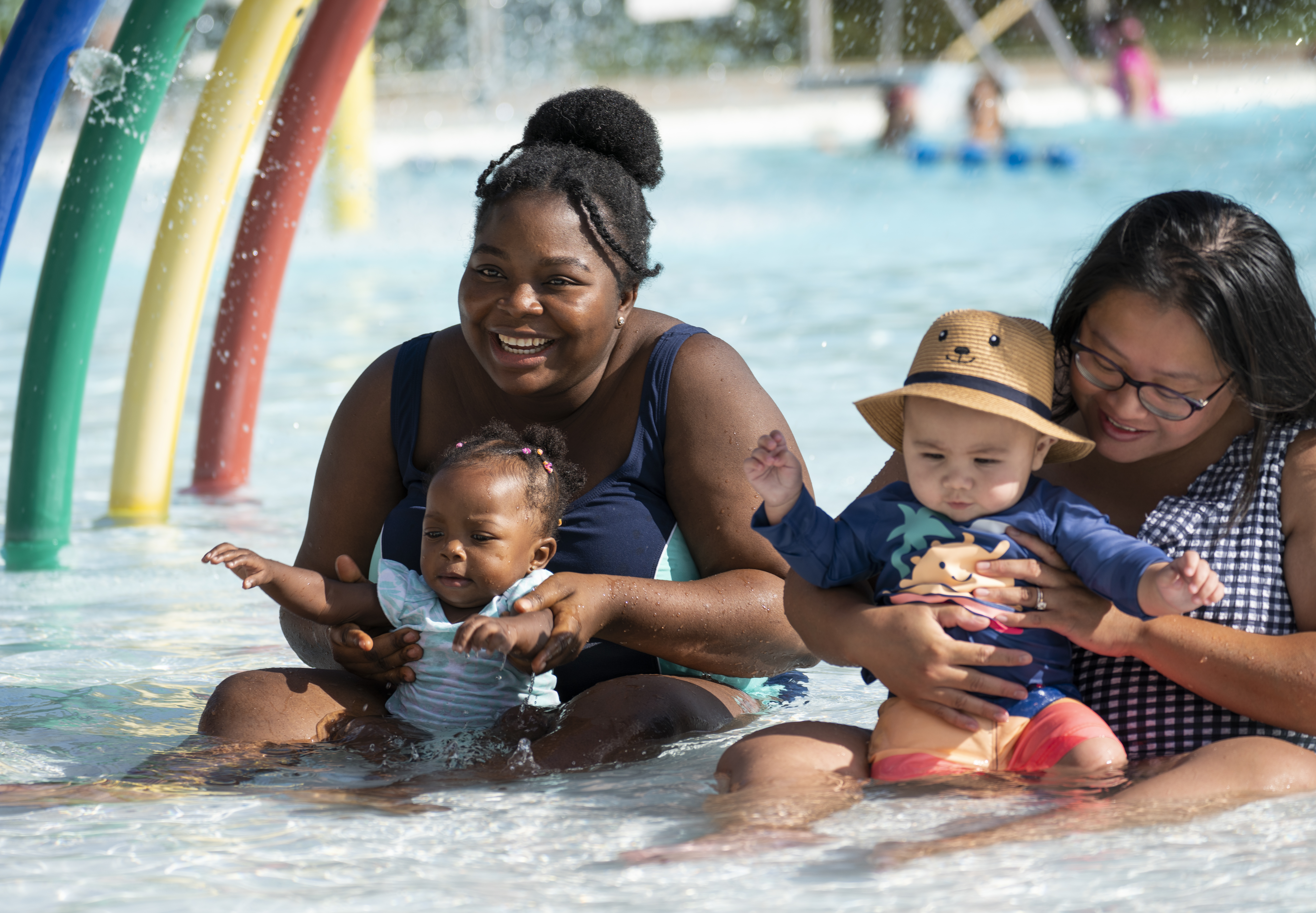 Two moms with their infants sitting in the water at the lake lisgar waterpark on a bright sunny day