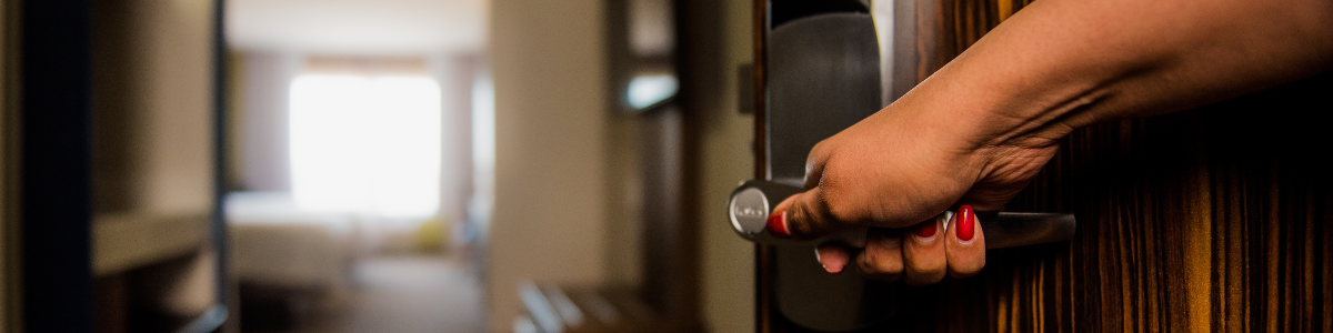 A hand on the hotel room door, opens it to reveal a clean, cozy and bright hotel room